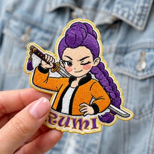 May include: An embroidered patch featuring a character with purple hair in a long braid, wearing an orange jacket over a white shirt. The character holds a katana over their shoulder and winks. The name RUMI is stitched in purple at the bottom.