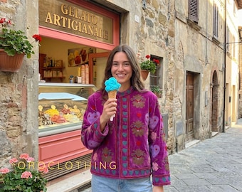 May include: A woman wearing a magenta jacket with embroidered heart and floral designs, holding a blue ice cream cone in front of a gelato shop. The shop sign reads "Gelato Artigianale". The text "OROCLOTHING" is visible at the bottom.