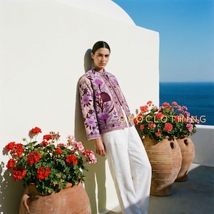 Puede incluir: Una mujer con una chaqueta rosa claro con bordados florales morados y pantalones blancos. La chaqueta tiene cuello mandarín y cierre de botones. Flores rojas en macetas de terracota en primer plano, con el mar al fondo.