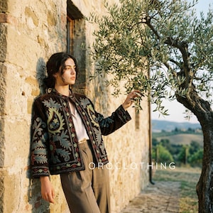 May include: A brown embroidered jacket with floral patterns in green, blue, and red. The jacket is worn over a white blouse and brown trousers. The text "OROCLOTHING" is visible. The jacket has a mandarin collar.