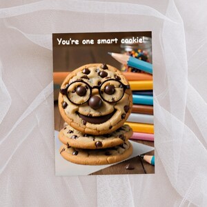 May include: A greeting card featuring a stack of three chocolate chip cookies. The top cookie has dark chocolate chip eyes, a round chocolate nose, a chocolate smile, and black glasses. The text reads, You're one smart cookie! Coloured pencils are visible.