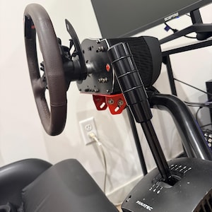 May include: A racing simulator setup featuring a brown leather steering wheel, a black gear shift, and a black motor. The setup includes a red mounting bracket and a black seat. The word "FANATEC" is visible on the gear shift.