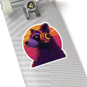 May include: A sticker featuring a purple raccoon wearing orange headphones, set against a retro sunset design. The raccoon is looking to the left. The sticker is on a skateboard.