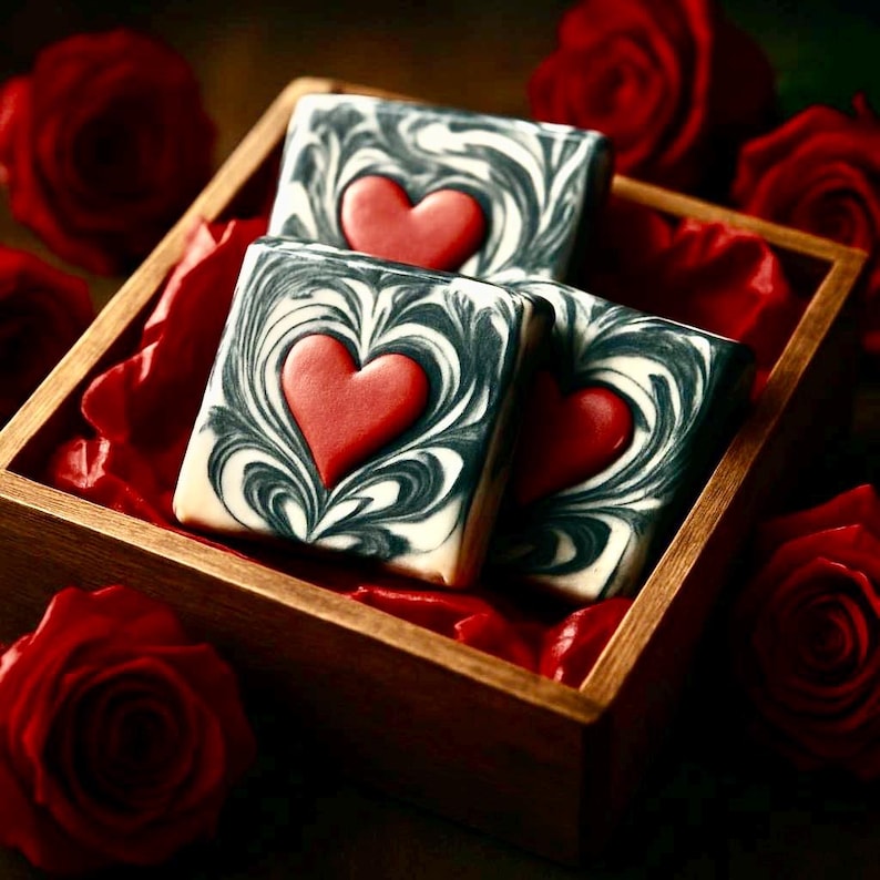 May include: Three square soaps with a red heart design are displayed in a wooden box lined with red tissue paper. The soaps have a black and white swirl pattern. Red roses are scattered around the box.