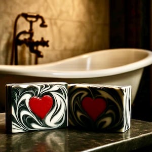 May include: Two handmade soaps with a red heart design. The soaps have a black and white swirl pattern. The background includes a white bathtub and vintage faucet.