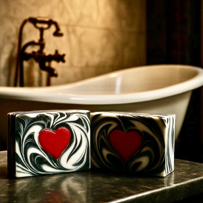May include: Two handmade soaps with a red heart design. The soaps have a black and white swirl pattern. The background includes a white bathtub and vintage faucet.