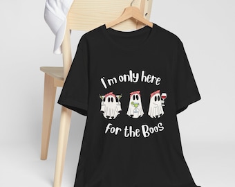 May include: Black t-shirt with the phrase "I'm only here for the Boos" in white. The shirt features three cartoon ghosts holding cocktails. A white baseball cap rests on a wooden chair in the background.