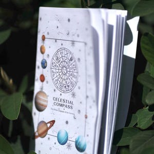 Könnte beinhalten: Ein weißes Softcover-Buch mit dem Titel Celestial Compass: The Divine Language of the Universe. Das Cover zeigt ein zentrales kreisförmiges Tierkreiszeichen, umgeben von Illustrationen von Planeten, Sternen und Sternbildern in Blau- und Orangetönen.