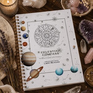 Könnte beinhalten: Ein spiralgebundenes Notizbuch mit weißem Einband, das ein schwarzes Tierkreiszeichen-Rad, himmlische Illustrationen und den Text "CELESTIAL COMPASS The Divine Language of the Universe. CURATED WITH LOVE BY LAINI REAGAN" zeigt. Der Einband enthält bunte Planeten.
