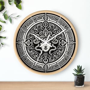 May include: Round wall clock with a light wood frame. The clock face features a black and white Medusa design. The clock hands are white and the numbers are indicated by lines. The clock is on a wooden surface.