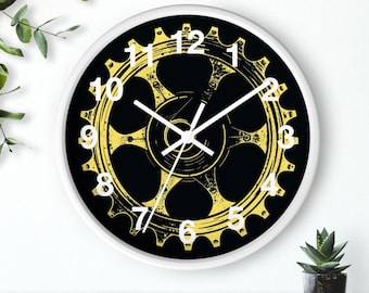 Puede incluir: Un reloj de pared redondo con marco blanco. La esfera negra presenta un diseño de engranaje amarillo y números blancos. Las manecillas del reloj son blancas. El reloj está sobre una superficie de madera.