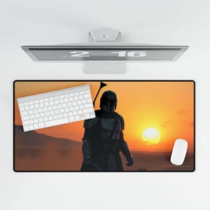 Op de afbeelding: Een rechthoekige muismat met een zonsondergangscène en een silhouet in harnas. Een wit toetsenbord en muis liggen op de mat. De afbeelding staat tegen een witte achtergrond, met een computermonitor erboven.