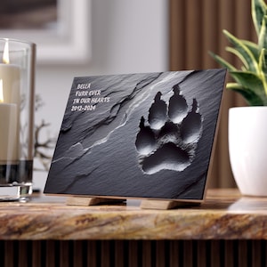 May include: A black slate pet memorial plaque with a paw print design. The plaque features the text "Bella Furr Ever In Our Hearts 2012-2024" in white lettering. The plaque is supported by two small wooden stands.