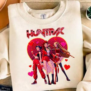 May include: Cream-colored sweatshirt featuring a graphic of three stylized figures in red outfits, set against a pink heart backdrop with the word "HUNTRAX" above. The design includes hearts and a dotted pattern.