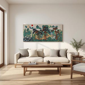 Op de afbeelding: Een rechthoekig landschapsschilderij met een berglandschap met bomen, water en een rode zon. Het kunstwerk is gemonteerd boven een crèmekleurige bank met grijze en witte kussens. Een houten salontafel en stoel staan voor de bank.