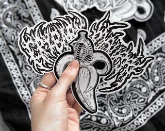 May include: Black and white sticker featuring a banana with a skeleton torso emerging from the peel. The sticker is held in a hand, with other similar stickers in the background. The background is a black and white paisley pattern.