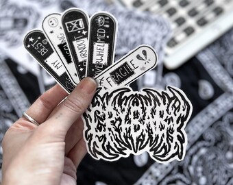 May include: A hand holding a collection of black and white stickers. The stickers include phrases like "Best," "My," "Burnout," "Overwhelmed," and "Fragile." A large, abstract black and white sticker is also visible.