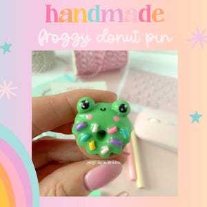 May include: A handmade Frogggy donut pin. The pin features a green frog with black eyes and a smiling mouth, on a donut with colourful sprinkles. The background is a pastel gradient with text that reads "handmade Frogggy donut pin".