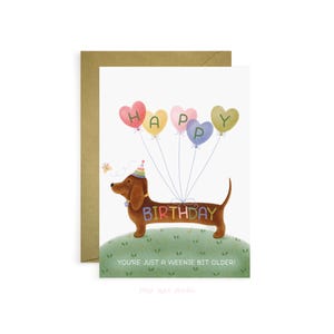 May include: A birthday card with a brown dachshund holding balloons spelling "HAPPY" and "BIRTHDAY". The card reads "YOU'RE JUST A WEENIE BIT OLDER!". The card is on a white background with a brown envelope.