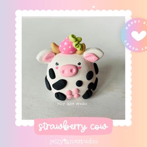 May include: A handcrafted strawberry cow figurine. The white cow has black spots, pink ears, and a pink nose. A pink strawberry with green leaves sits atop its head. The text "strawberry cow" is displayed below the figurine.
