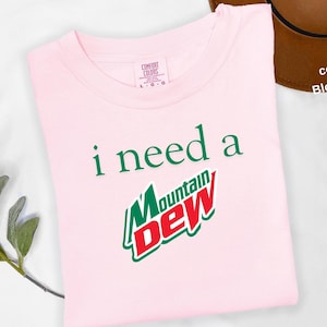 May include: A light pink t-shirt with the text "i need a Mountain Dew" in green and red. The Mountain Dew logo is prominently displayed. The shirt is a Comfort Colors brand, size L-G.