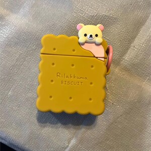 May include: Yellow AirPod case shaped like a biscuit, with a cute bear character peeking over the top. The case has the words "Rilakkuma BISCUIT" printed on it. A pink loop is attached to the side.