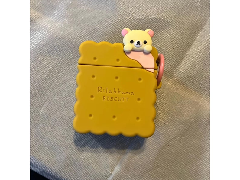 May include: Yellow AirPod case shaped like a biscuit, with a cute bear character peeking over the top. The case has the words "Rilakkuma BISCUIT" printed on it. A pink loop is attached to the side.