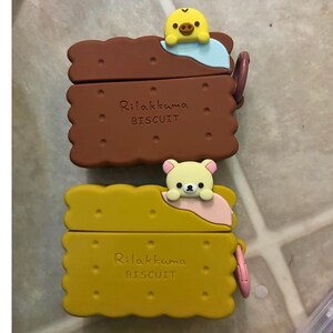 May include: Two Rilakkuma Biscuit-shaped cases, one brown and one yellow, each with a cartoon character on top. The brown case has a yellow chick, and the yellow case has a bear. The text "Rilakkuma BISCUIT" is printed on each case.