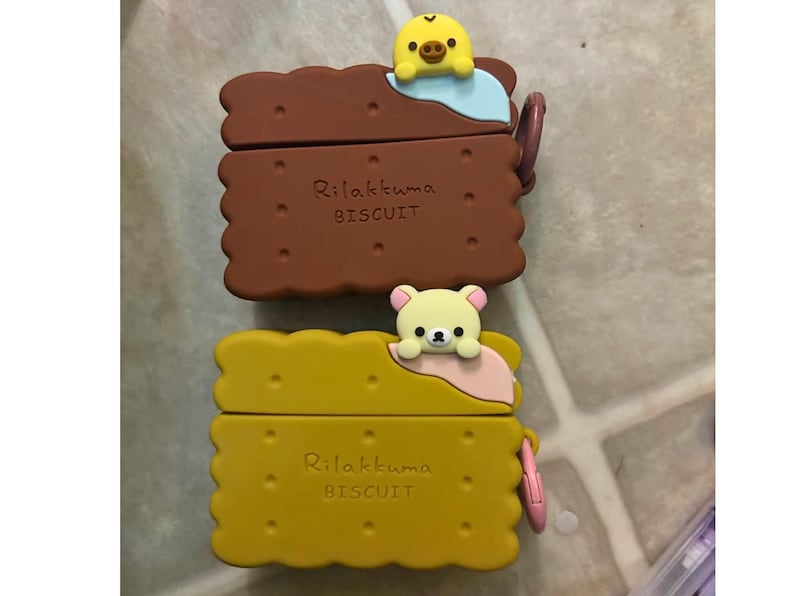 May include: Two Rilakkuma Biscuit-shaped cases, one brown and one yellow, each with a cartoon character on top. The brown case has a yellow chick, and the yellow case has a bear. The text "Rilakkuma BISCUIT" is printed on each case.