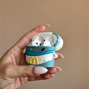 May include: A blue, white, and yellow silicone case for wireless earbuds. The case is designed to resemble a fast-food cup, with a yellow "M" logo. The earbuds are white. The case is held open.