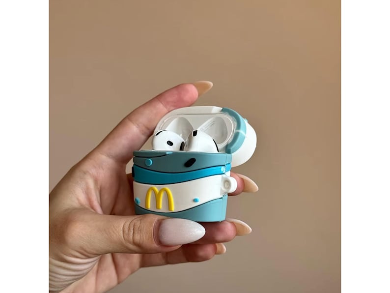 May include: A blue, white, and yellow silicone case for wireless earbuds. The case is designed to resemble a fast-food cup, with a yellow "M" logo. The earbuds are white. The case is held open.