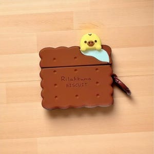 May include: A brown biscuit-shaped case with the text "Rilakkuma BISCUIT" and a yellow and light blue character on top. The case has a dark brown carabiner clip attached to the side.