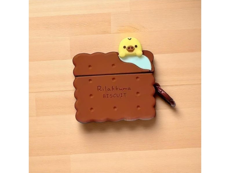 May include: A brown biscuit-shaped case with the text "Rilakkuma BISCUIT" and a yellow and light blue character on top. The case has a dark brown carabiner clip attached to the side.