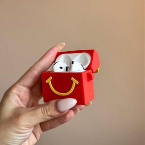 May include: Red AirPods case with a yellow smiling face design. The case is open, revealing white AirPods. The case has a rectangular shape with rounded edges. The background is a solid beige color.