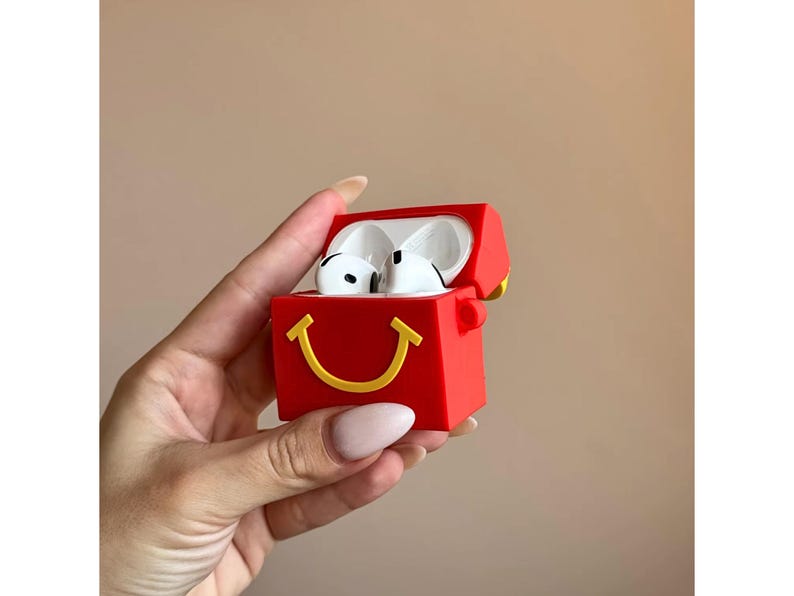 May include: Red AirPods case with a yellow smiling face design. The case is open, revealing white AirPods. The case has a rectangular shape with rounded edges. The background is a solid beige color.