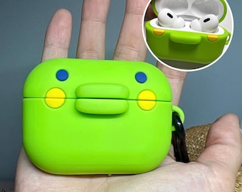 May include: A bright green silicone case for wireless earbuds, designed with a cartoon frog face. The case features blue and yellow accents, a hinged lid, and a small black strap. A smaller inset image shows the case open with white earbuds inside.