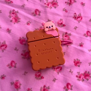 May include: A brown biscuit-shaped case for electronic devices, featuring the text "Rilakkuma BISCUIT" and a small pink bear figure on top. The case has a pink loop and is set against a pink floral background.