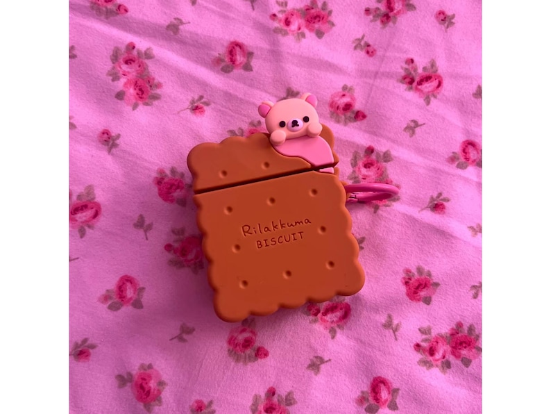 May include: A brown biscuit-shaped case for electronic devices, featuring the text "Rilakkuma BISCUIT" and a small pink bear figure on top. The case has a pink loop and is set against a pink floral background.