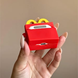 May include: A red and yellow silicone case for wireless earbuds, designed to resemble a fast-food french fries container. The case has a rectangular shape with a yellow arch on top and a charging port opening at the bottom.