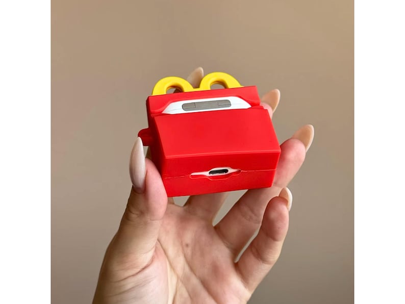 May include: A red and yellow silicone case for wireless earbuds, designed to resemble a fast-food french fries container. The case has a rectangular shape with a yellow arch on top and a charging port opening at the bottom.