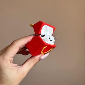 May include: Red AirPods case shaped like a fast food container, with a yellow arch logo. The case is open, revealing white AirPods inside. The case has a yellow hinge and is held by a hand.