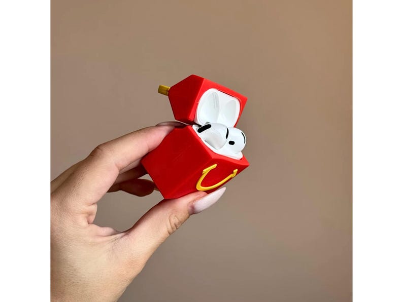 May include: Red AirPods case shaped like a fast food container, with a yellow arch logo. The case is open, revealing white AirPods inside. The case has a yellow hinge and is held by a hand.