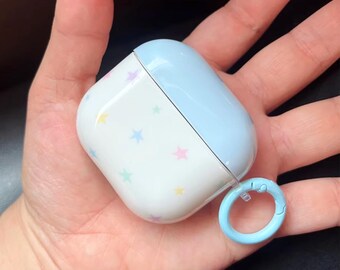 May include: A white and light blue AirPod case with a star pattern. The case has a light blue lid and a matching light blue carabiner clip. The case is held in an open hand.