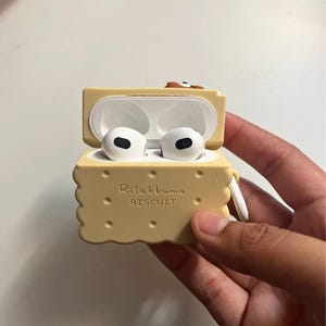 May include: A light yellow, biscuit-shaped AirPods case with the text "Rilakkuma BISCUIT" printed on the front. The case is open, revealing white AirPods inside. The case has a small loop for attaching to a keychain.