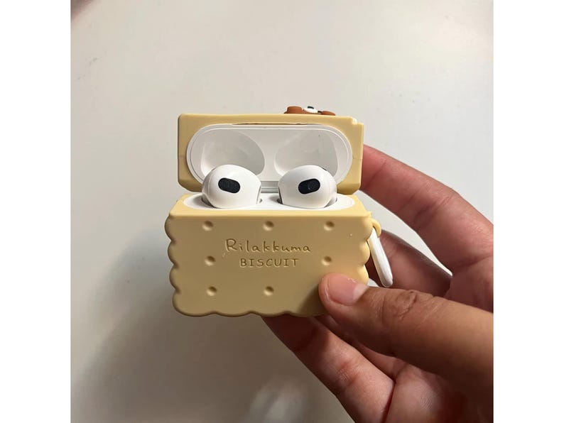 May include: A light yellow, biscuit-shaped AirPods case with the text "Rilakkuma BISCUIT" printed on the front. The case is open, revealing white AirPods inside. The case has a small loop for attaching to a keychain.