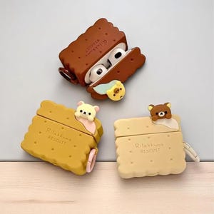 Può includere: Tre custodie per AirPods a forma di biscotto, ognuna con un personaggio di orso diverso. Le custodie sono marroni, gialle e beige, con la scritta "Rilakkuma Biscuit" stampata. Una custodia è aperta, rivelando gli AirPods bianchi.