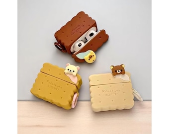 May include: Three AirPod cases in the shape of biscuits, each with a different bear character. The cases are brown, yellow, and beige, with the text "Rilakkuma Biscuit" printed on them. One case is open, revealing the white AirPods.