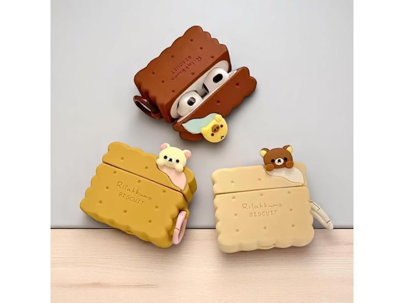 May include: Three AirPod cases in the shape of biscuits, each with a different bear character. The cases are brown, yellow, and beige, with the text "Rilakkuma Biscuit" printed on them. One case is open, revealing the white AirPods.