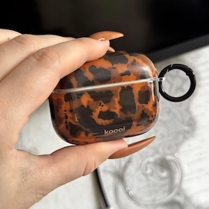 Może przedstawiać: Brązowo-czarne etui na AirPods z nadrukiem w panterkę z czarnym karabińczykiem. Na przodzie etui nadrukowano napis "koooi". Etui jest trzymane w dłoni.