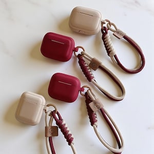 May include: AirPod cases in beige and burgundy with matching wrist straps. The cases are square-shaped and have a smooth finish. The straps are made of braided cord in beige and burgundy, with a small leather tag featuring a smiley face.